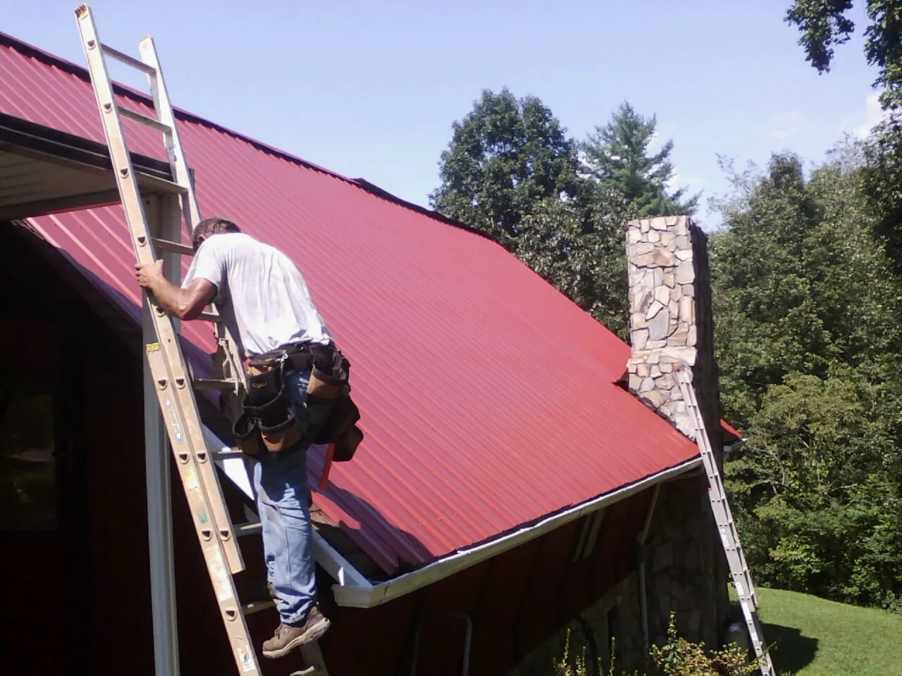 Roofer on a ladder beside a red metal roof and stone chimney — Soffit & Fascia Repair by Roofing Brothers in Reisterstown